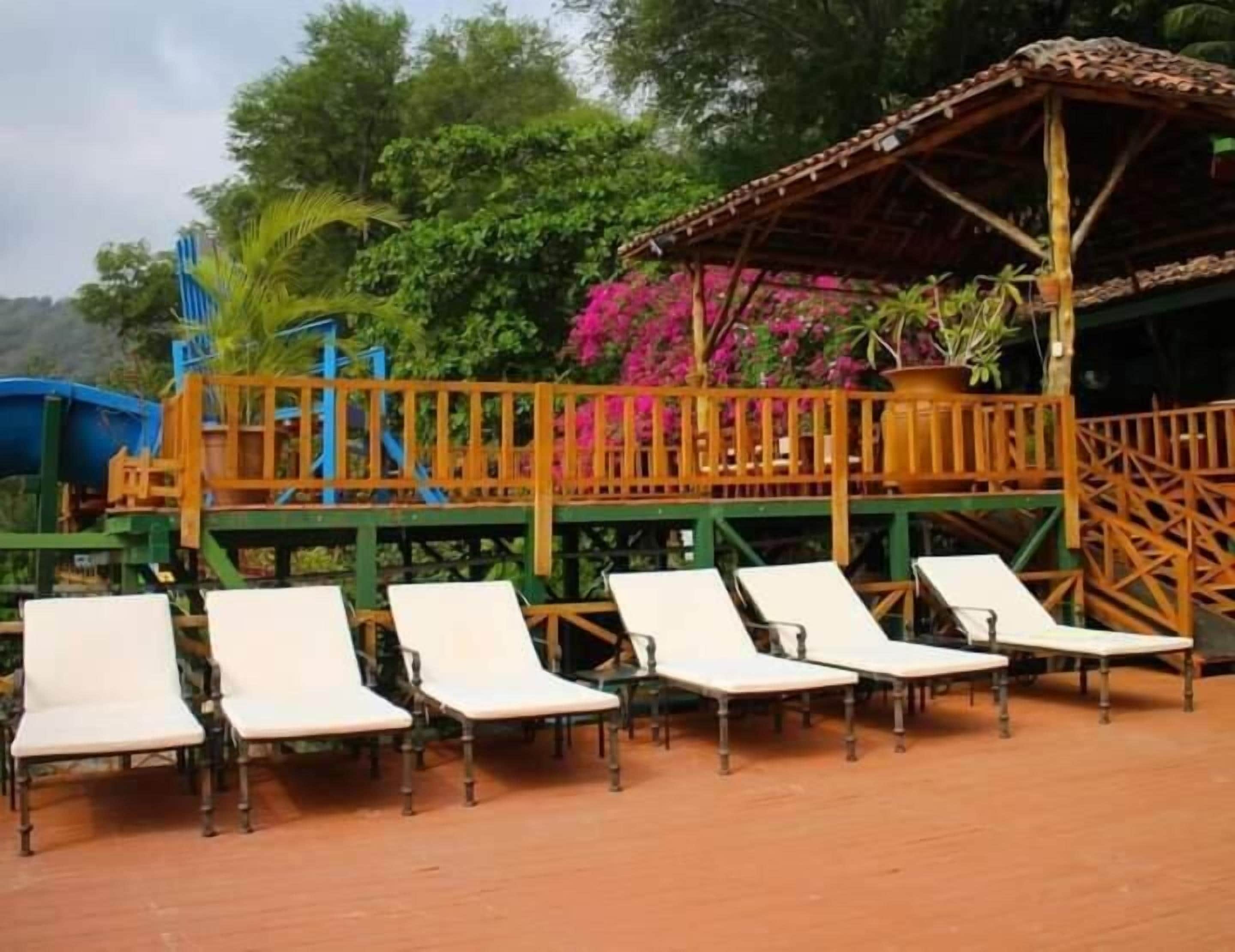 Een rij witte lounge fauteuils met metalen poten, een houten terras en een overdekt zitgedeelte met een metalen reling.