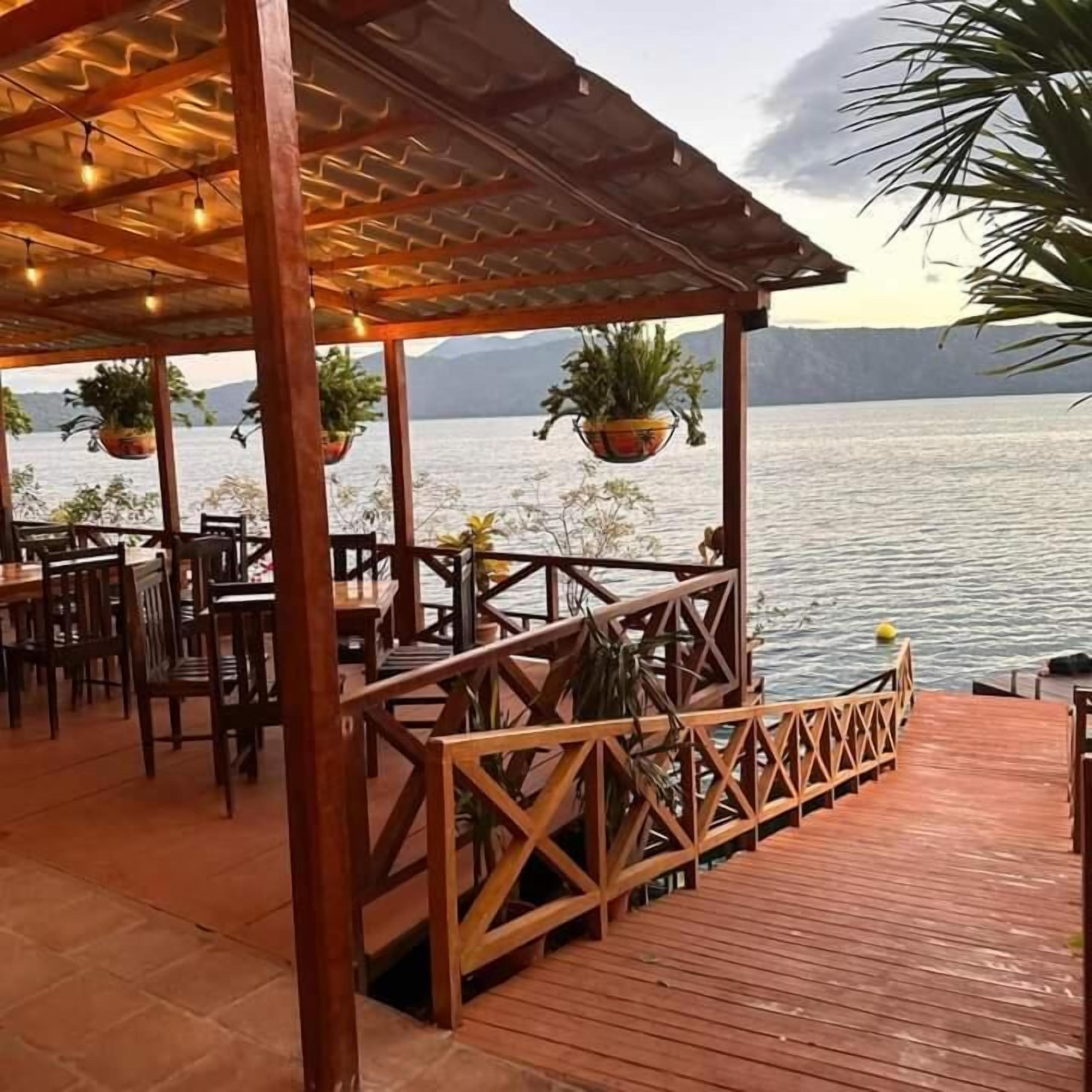 Une terrasse en bois avec des tables et des chaises donnant sur une étendue d’eau, agrémentée de jardinières suspendues et d’une rambarde.