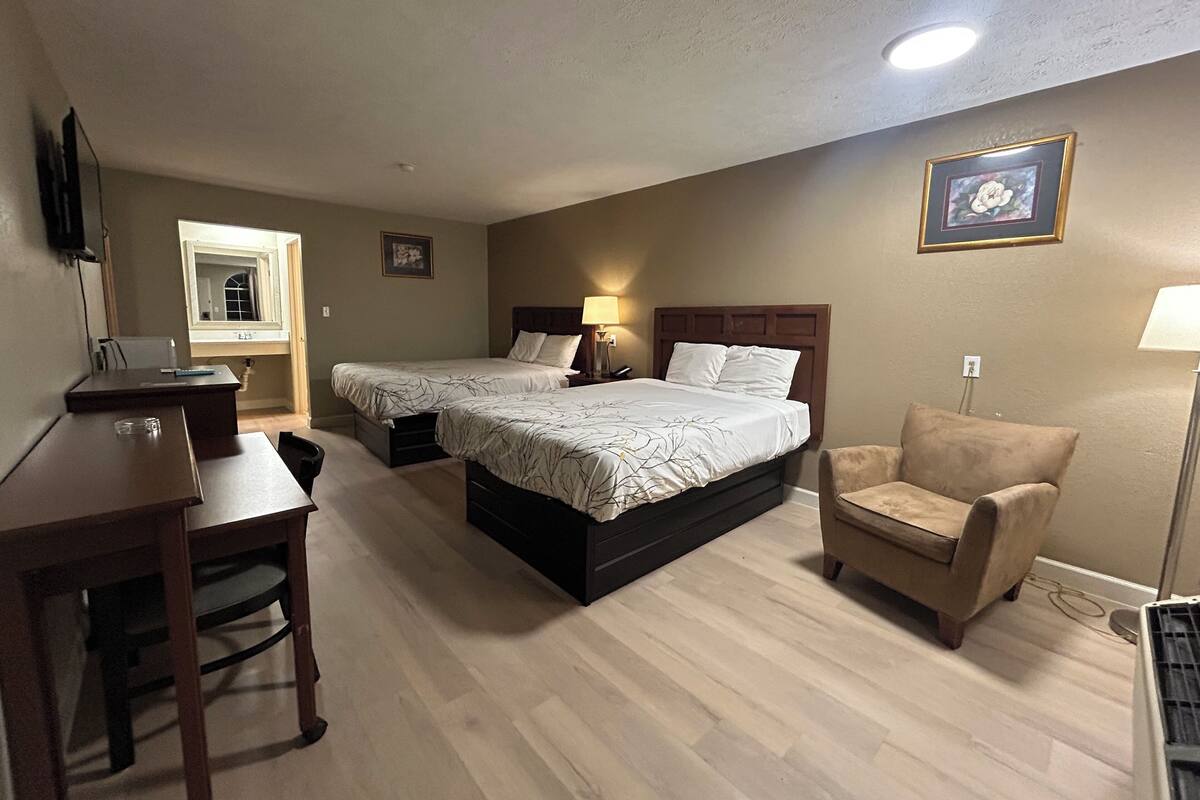 A hotel room with two beds, a desk, a chair, and a lamp.