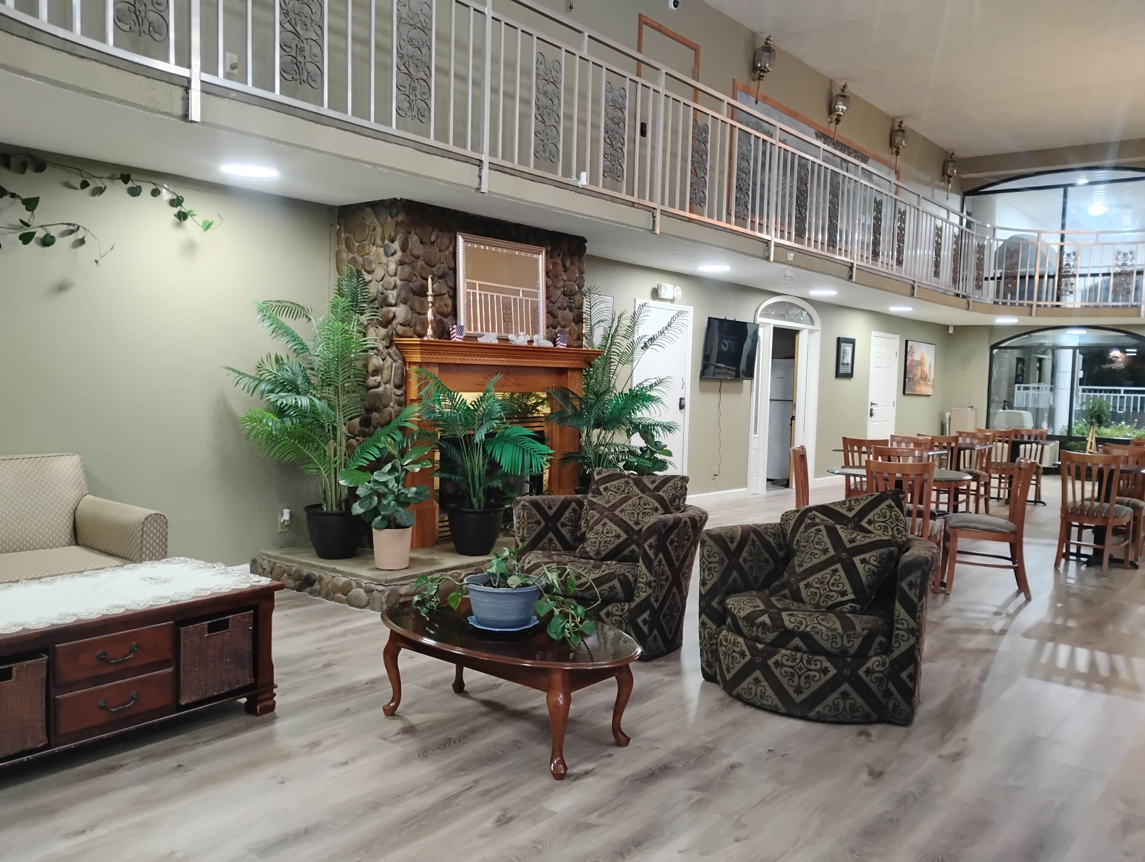 A spacious lobby with a high ceiling, a stone fireplace, multiple potted plants, patterned sofas, and a dining area with wooden chairs and tables.