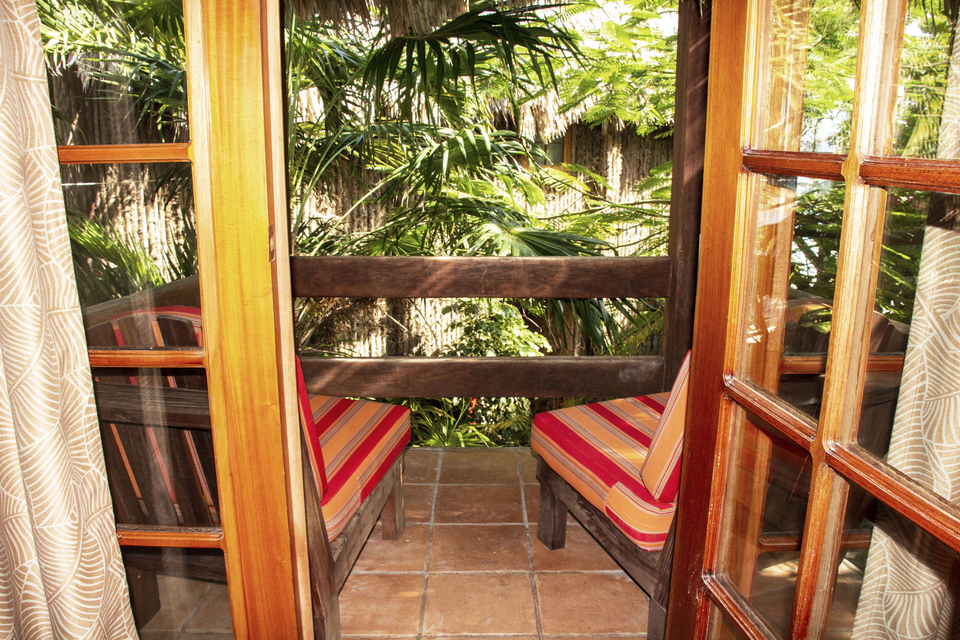 A wooden balcony with two cushioned benches, surrounded by glass walls and a view of lush greenery.