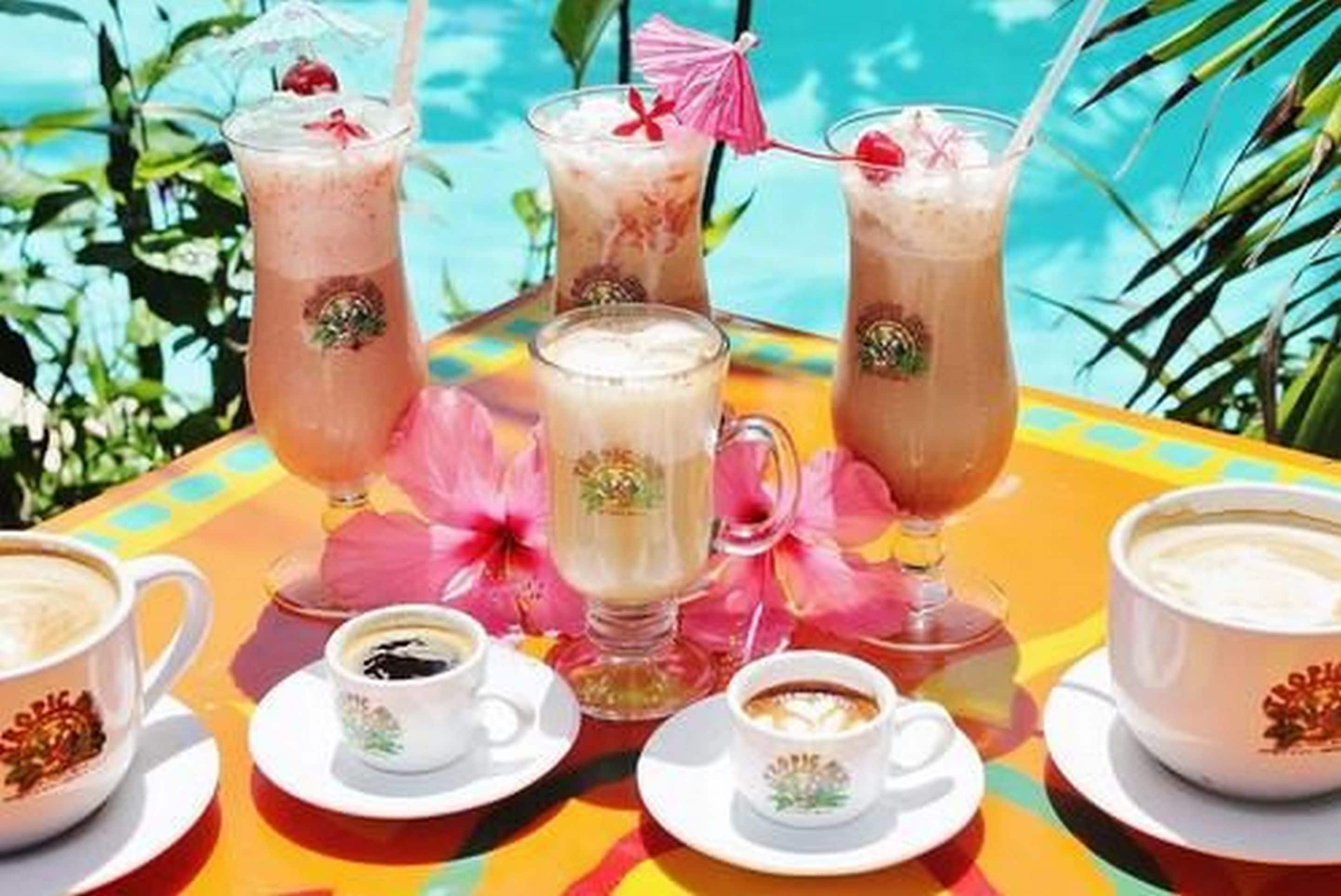 A table with various drinks and coffee cups, likely in a tropical setting with a pool in the background.