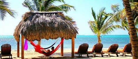 Una playa con una cabaña de techo de paja, sillas de descanso y una hamaca.