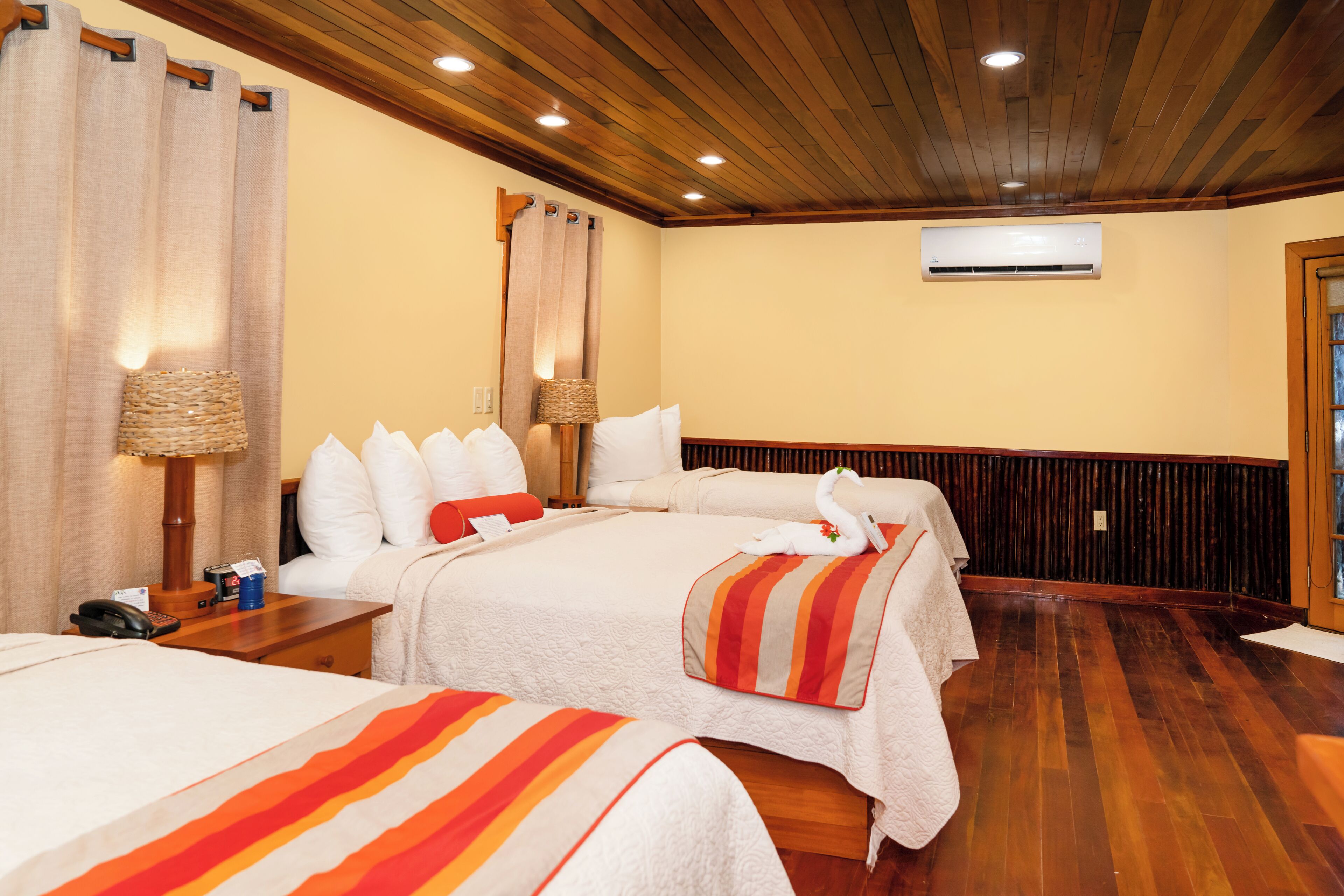 A hotel room with two beds, wooden flooring, a ceiling fan, an air conditioning unit, and a lamp on the nightstand.