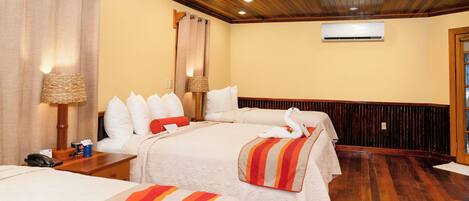 A hotel room with two beds, wooden flooring, a ceiling fan, an air conditioning unit, and a lamp on the nightstand.