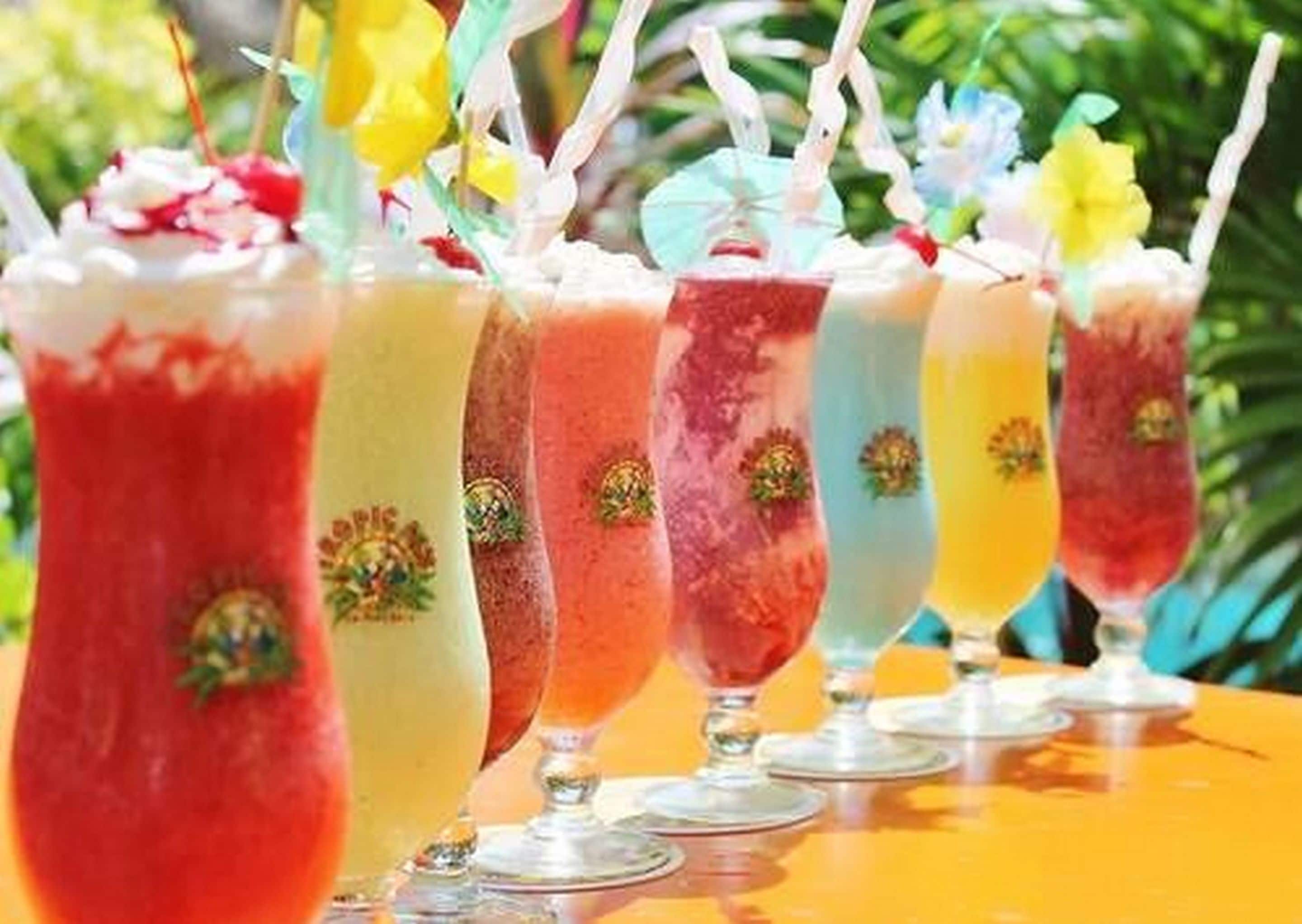 A row of colorful drinks in glasses with straws, placed on a table outdoors.