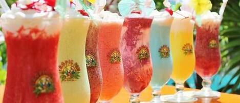 Una fila de bebidas coloridas en vasos con pajitas, sobre una mesa al aire libre.