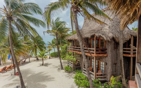 A beach house with a thatched roof, wooden balconies, and a sandy beach with palm trees.