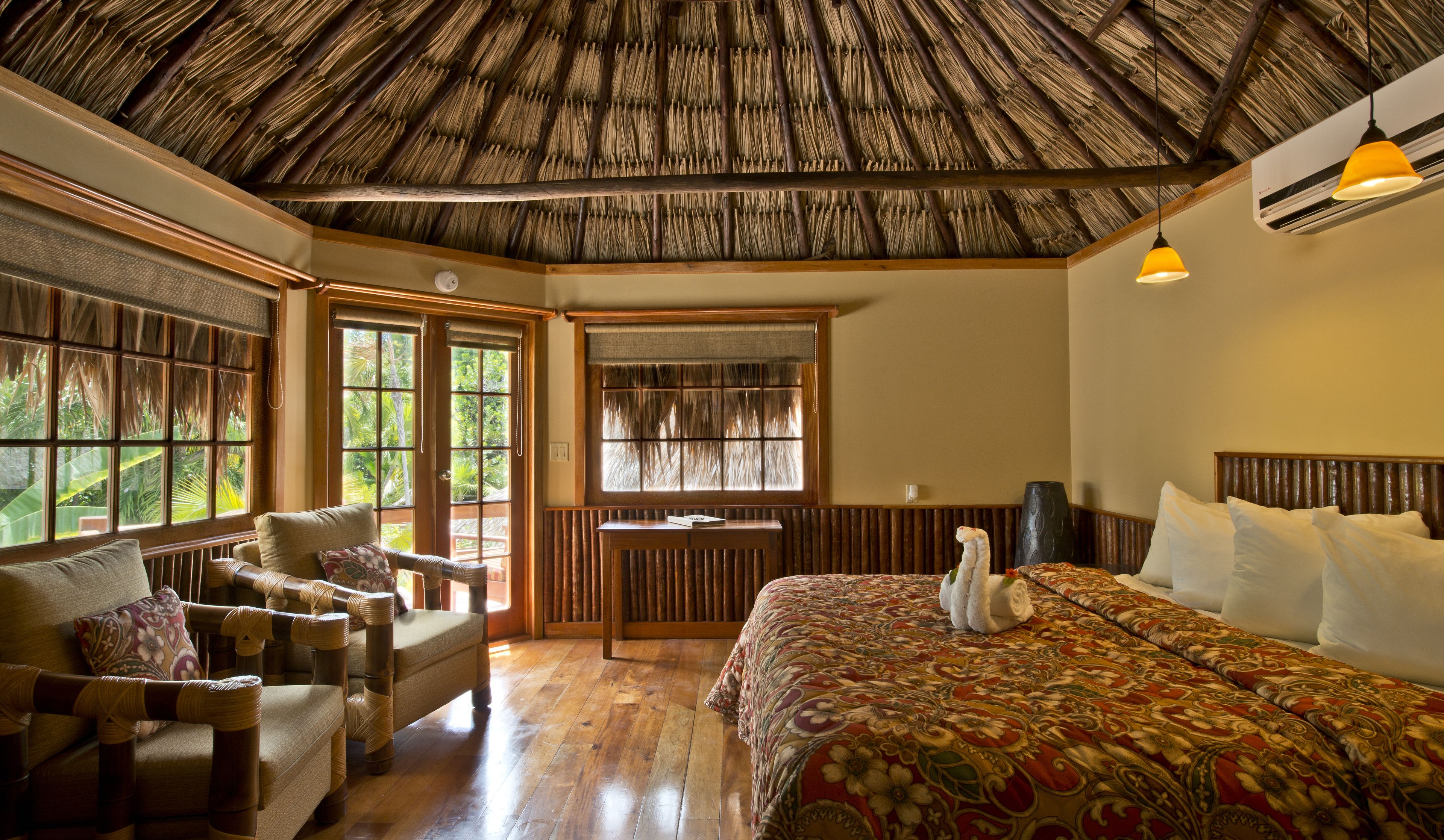 A room with a thatched roof, wooden furniture, a large bed with a floral bedspread, and large windows with a view of greenery.
