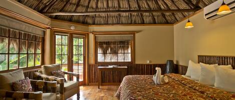 A room with a thatched roof, wooden furniture, a large bed with a floral bedspread, and large windows with a view of greenery.