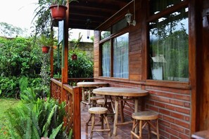 Un terrazzo in legno con tavoli e sgabelli, immerso nel verde e delimitato da un muro in mattoni.