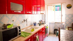 A kitchen with red cabinets, a microwave, a sink, and a window with blinds. - Next Stop Roma Affittacamere Casilino (Rome)