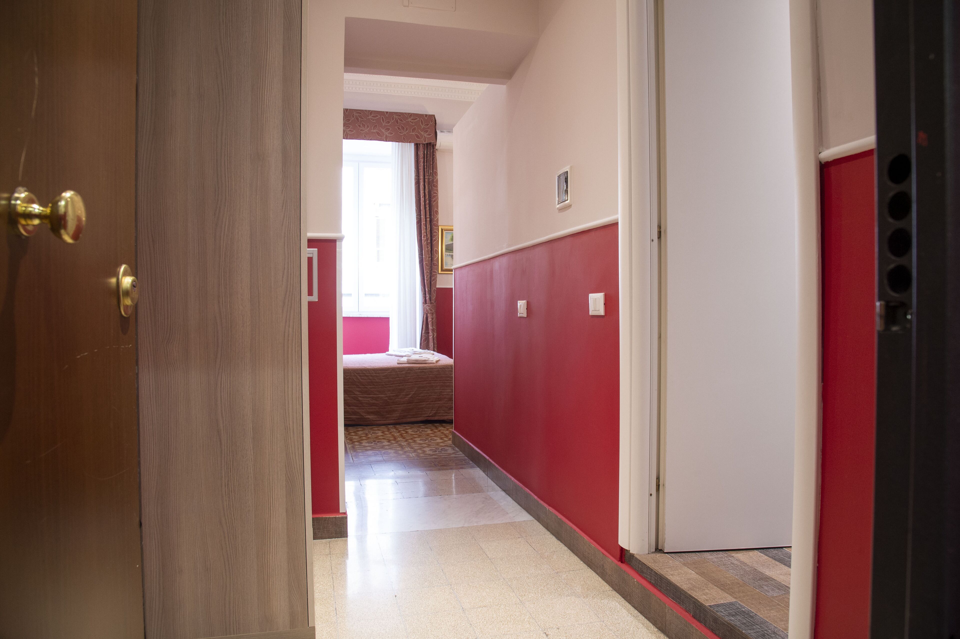 A hallway with a wooden door, a bed with a red wall, and a white door.
