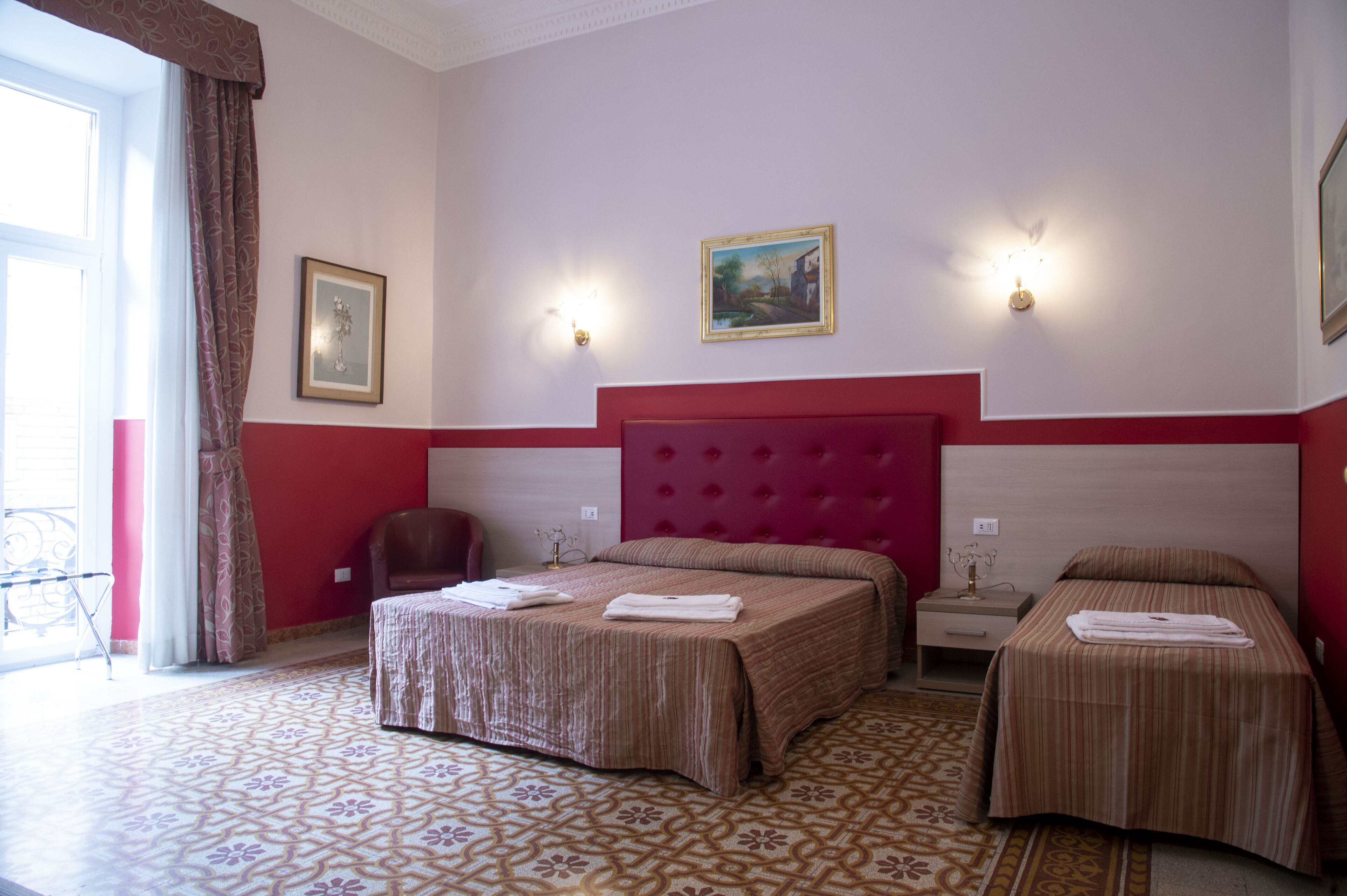 Une chambre d’hôtel avec deux lits, une tête de lit rouge, une petite table, une chaise et une fenêtre avec des rideaux.
