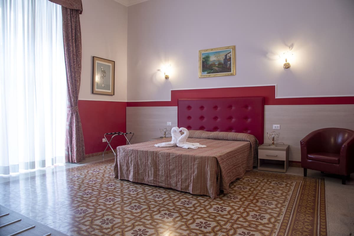 A hotel room with a large bed, a red headboard, two bedside tables, a red armchair, a patterned carpet, and a painting on the wall.