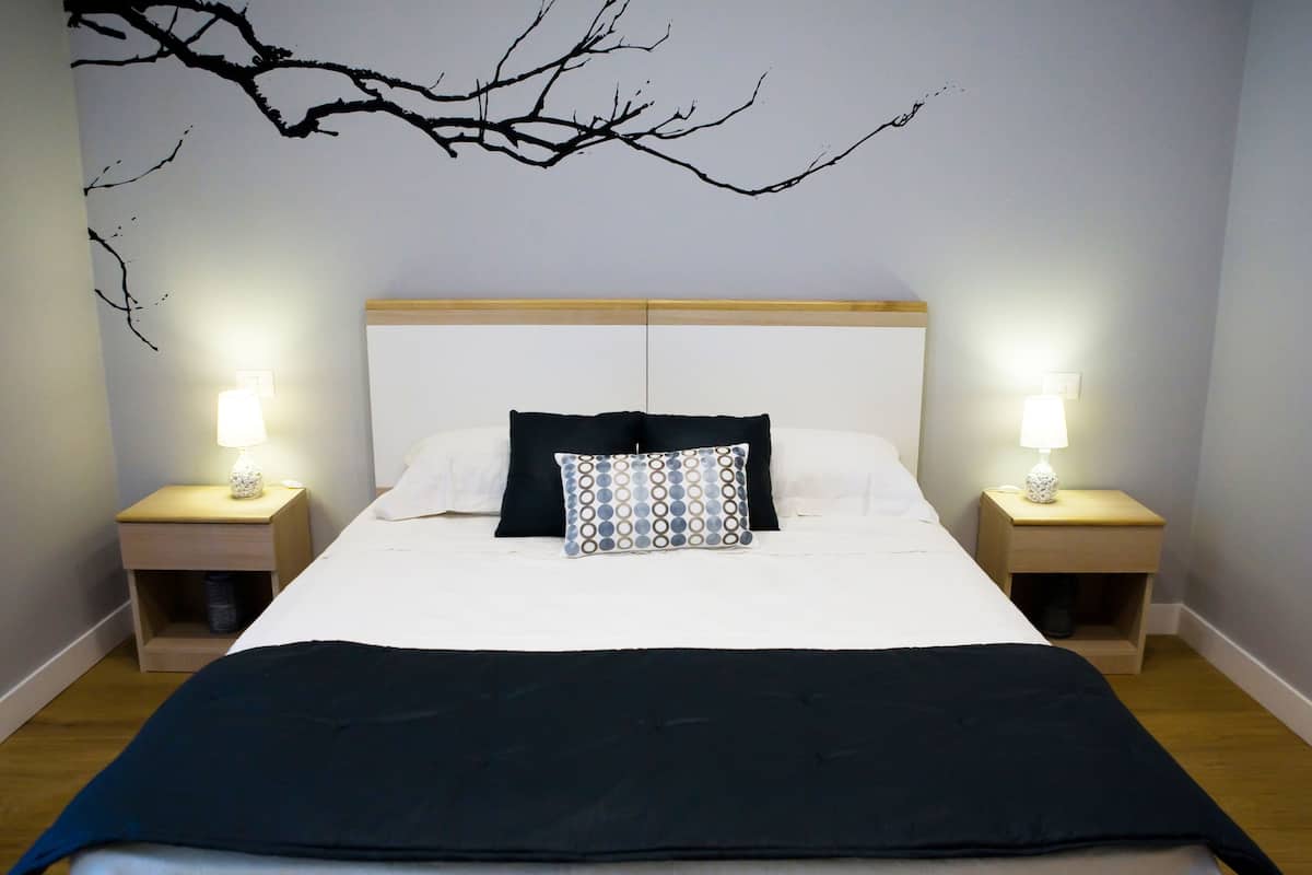 Una cama bien hecha con un edredón oscuro, dos mesitas de noche con lámparas, y una obra de arte en la pared que representa una rama de árbol.