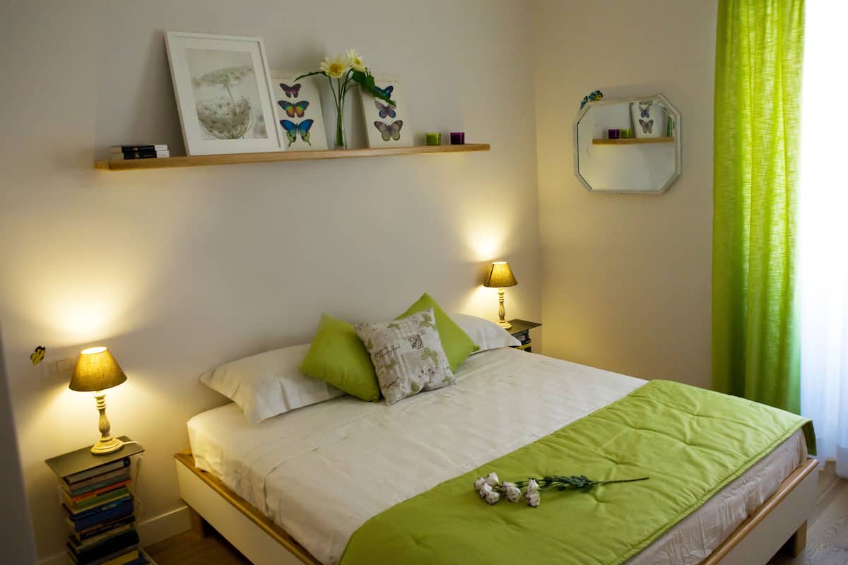 Un dormitorio ordenado con cama, lámparas de noche, mesita de noche con libros, un espejo y una estantería con objetos decorativos.