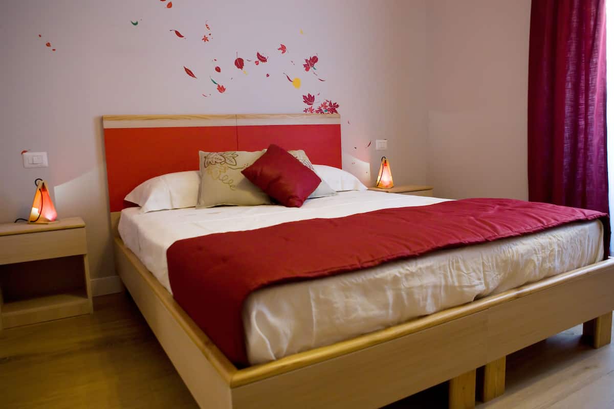 Una cama bien hecha con una colcha y almohadas rojas, un cabecero de madera y una pared decorada con motivos de hojas.