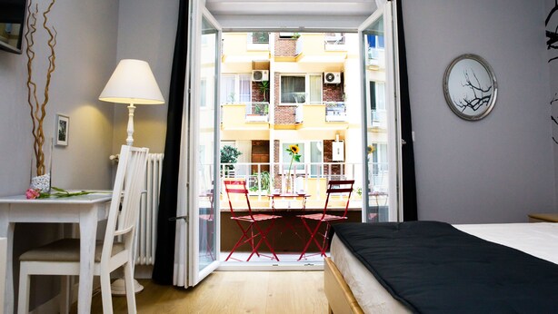 Une chambre comprenant un lit, un bureau avec une chaise, une table avec deux chaises, et donnant sur un balcon extérieur avec une balustrade.