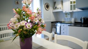 Un coin repas avec un vase violet rempli de fleurs posé sur une table, des chaises blanches et une cuisine en arrière-plan.