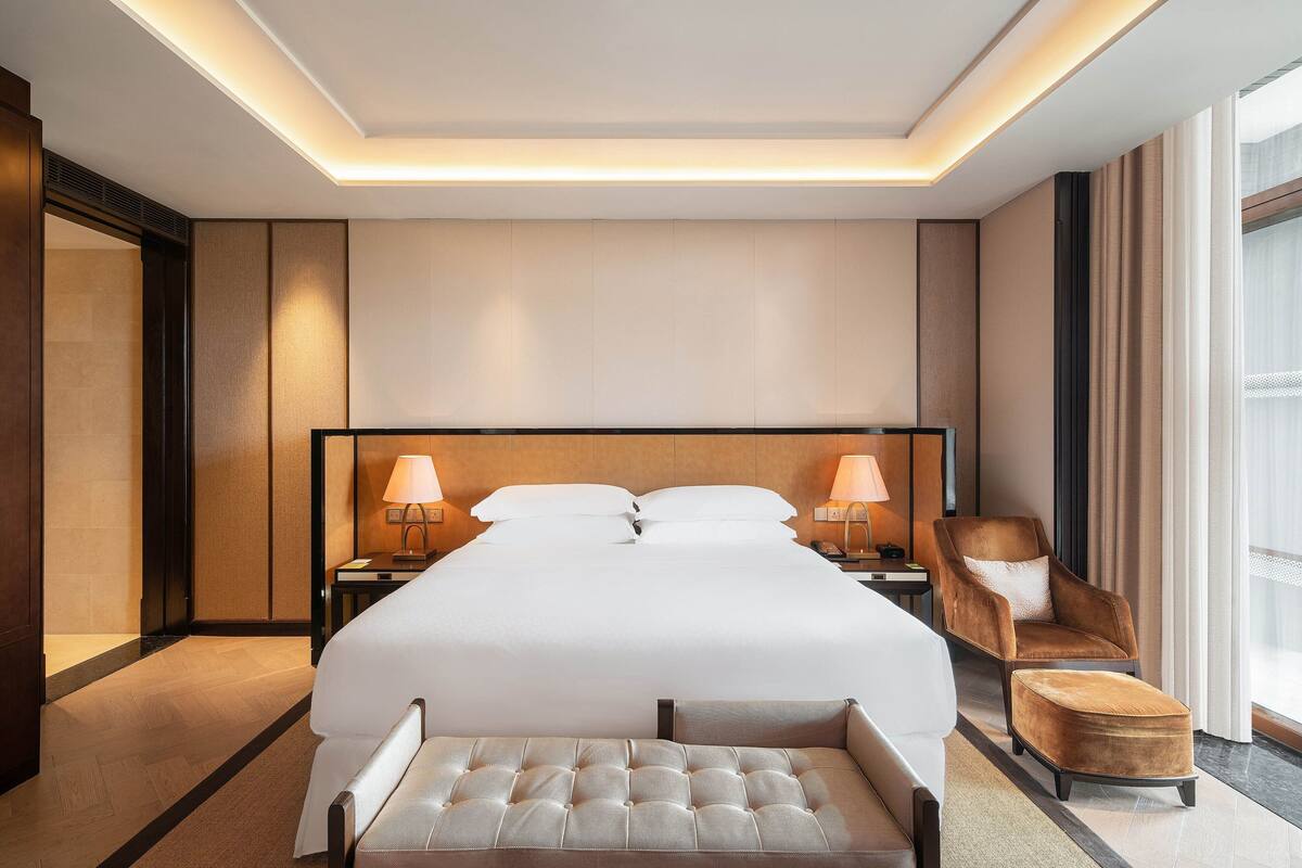 A modern hotel room with a large bed, two bedside lamps, a brown armchair, and a small ottoman.
