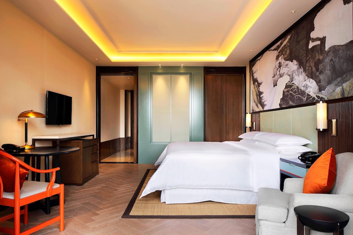 A hotel room with a bed, a desk, a chair, a TV, and a painting on the wall.