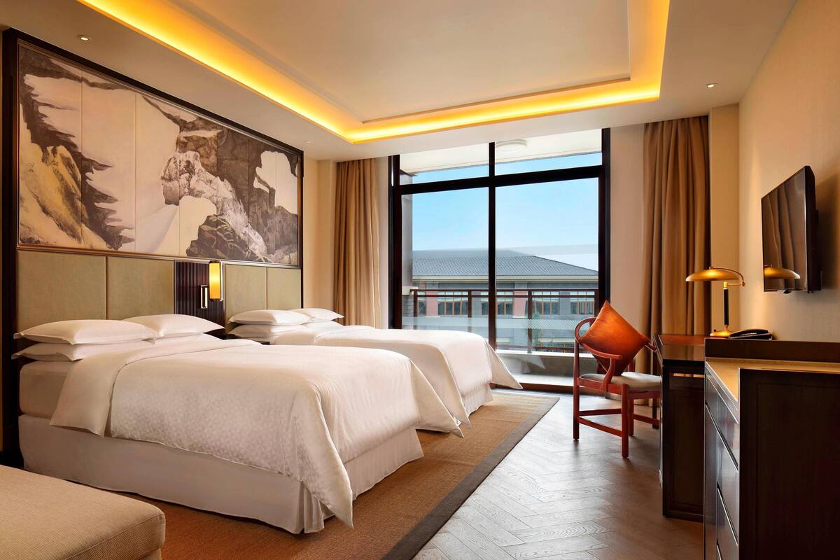A hotel room with two beds, a desk, a chair, a large window with a city view, and a mounted TV.