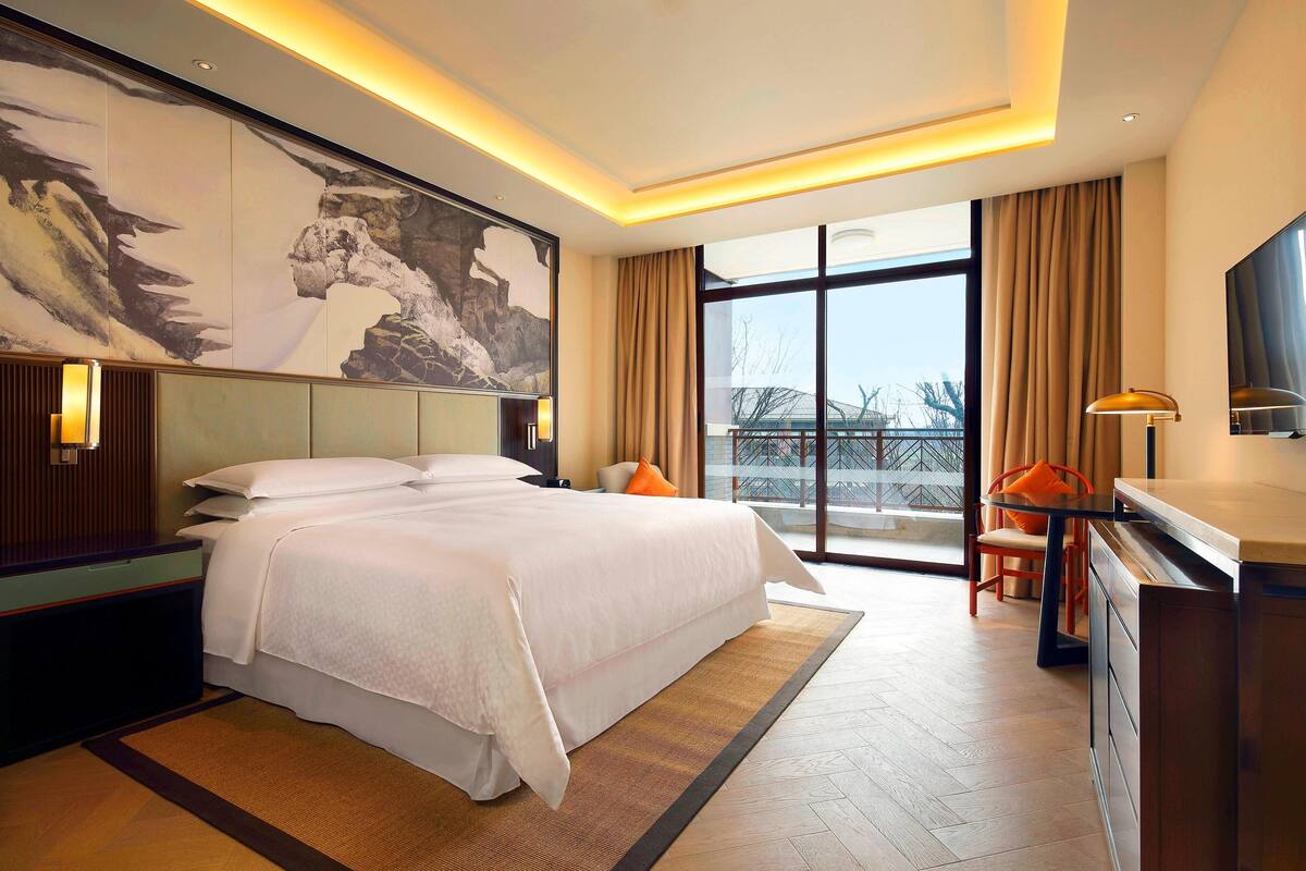 A hotel room with a large bed, a desk with a chair, a television, and a large window with curtains.