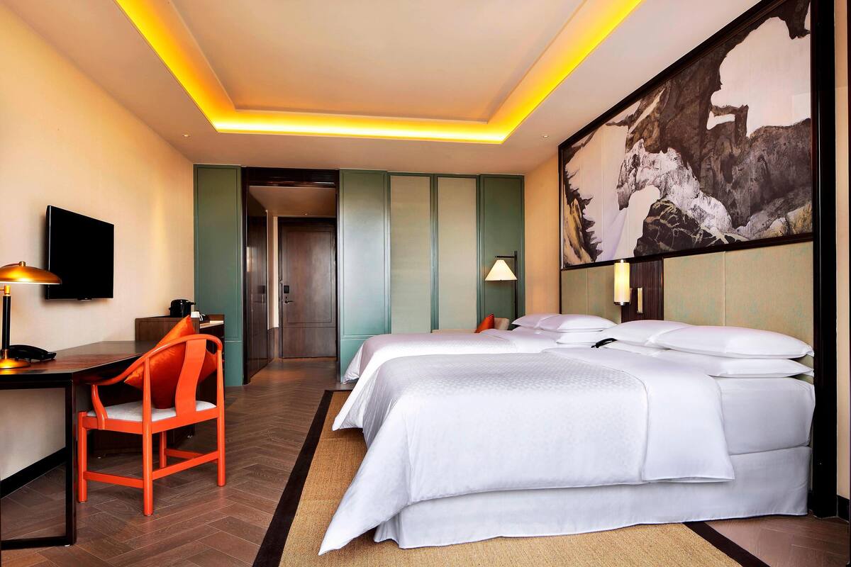 A hotel room with two beds, a desk, a chair, a TV, and a large painting on the wall.