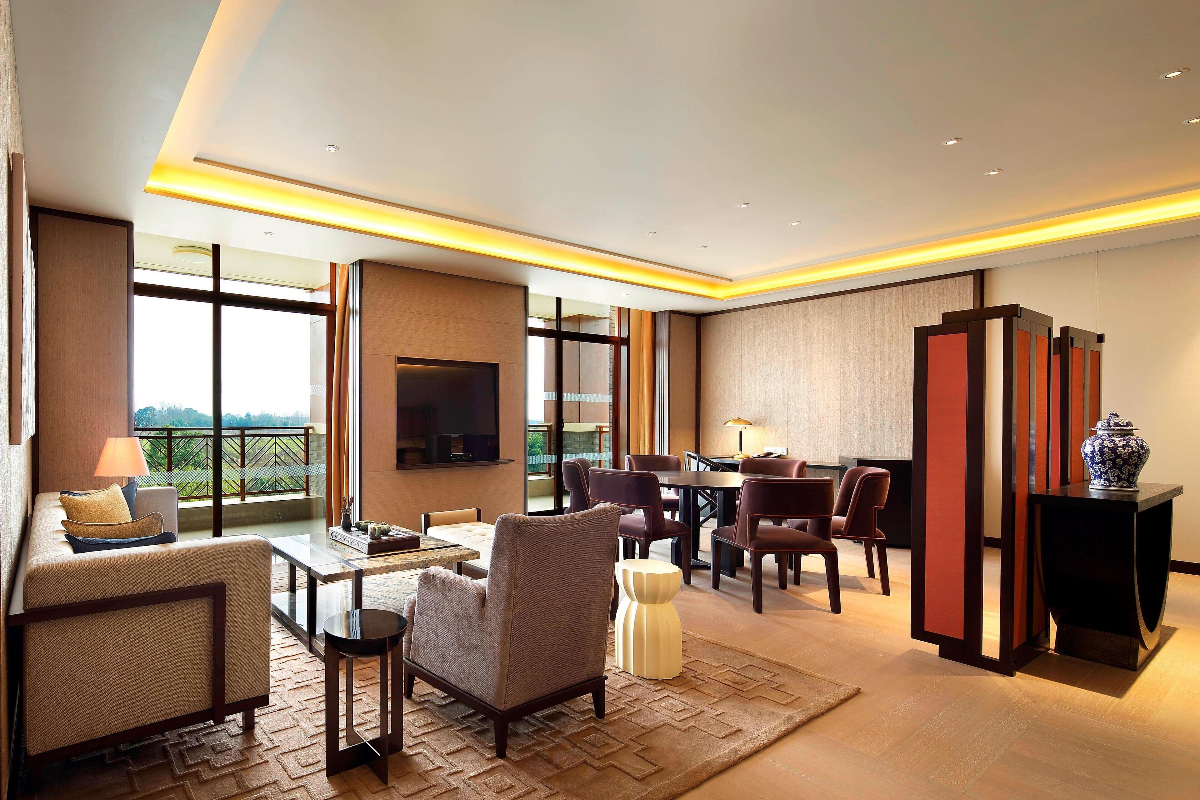 A spacious living area with a sofa, armchairs, a coffee table, and a dining table with chairs. A flat-screen TV is mounted on the wall, and there is a large window providing a view of the outdoors.