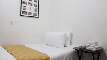 Ein Hotelzimmer mit einem Einzelbett, weißer Bettwäsche, einer gelben Tagesdecke, einem kleinen Tisch mit Handtüchern und einer gerahmten Fotocollage an der Wand.