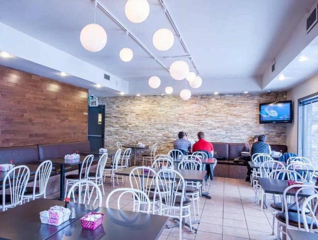Interior de restaurante con mesas altas, sillas blancas, pared de piedra y televisor de pantalla plana montado en la pared.
