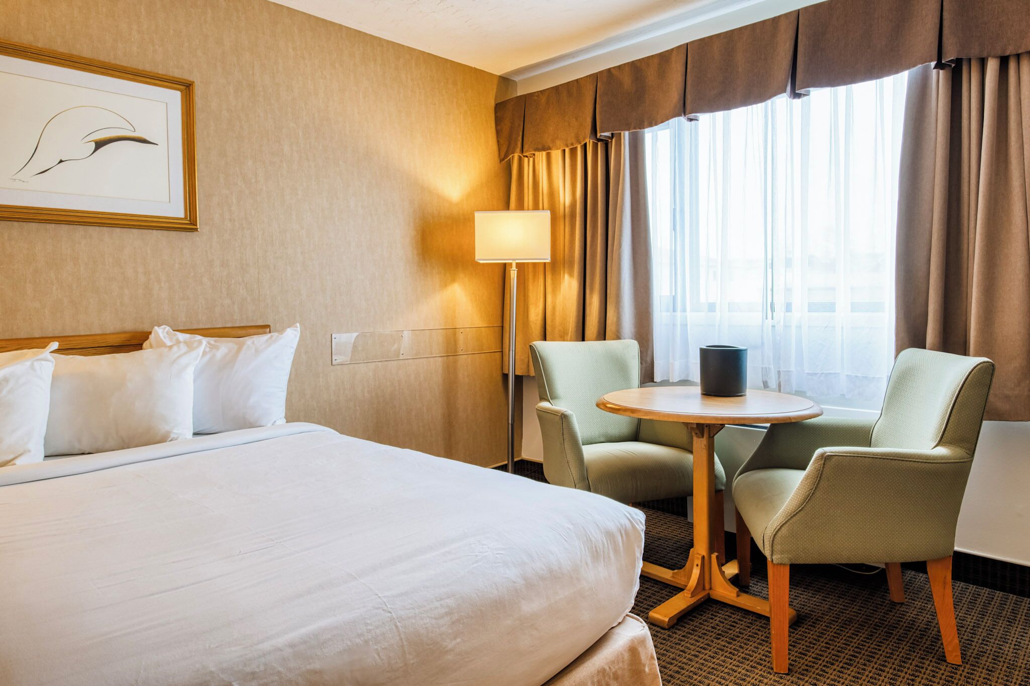 Una habitación de hotel con una cama grande, dos sillones, una mesita redonda, una lámpara y un ventanal con cortinas.