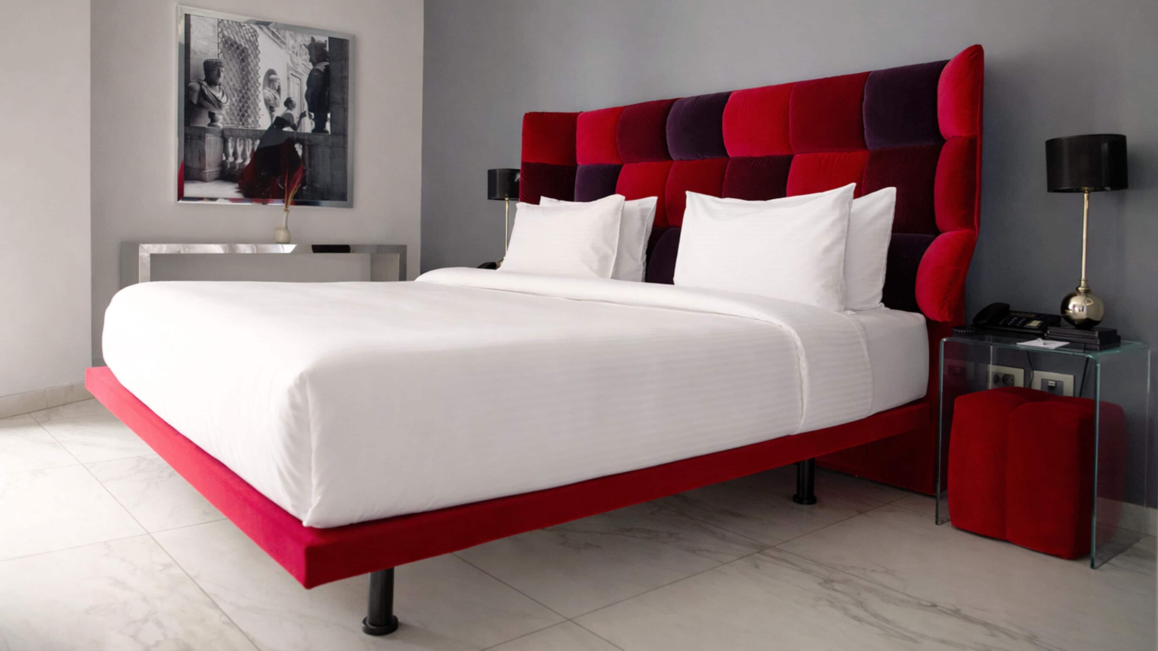 A modern bedroom with a red upholstered headboard, white bedding, and a glass bedside table with a lamp.