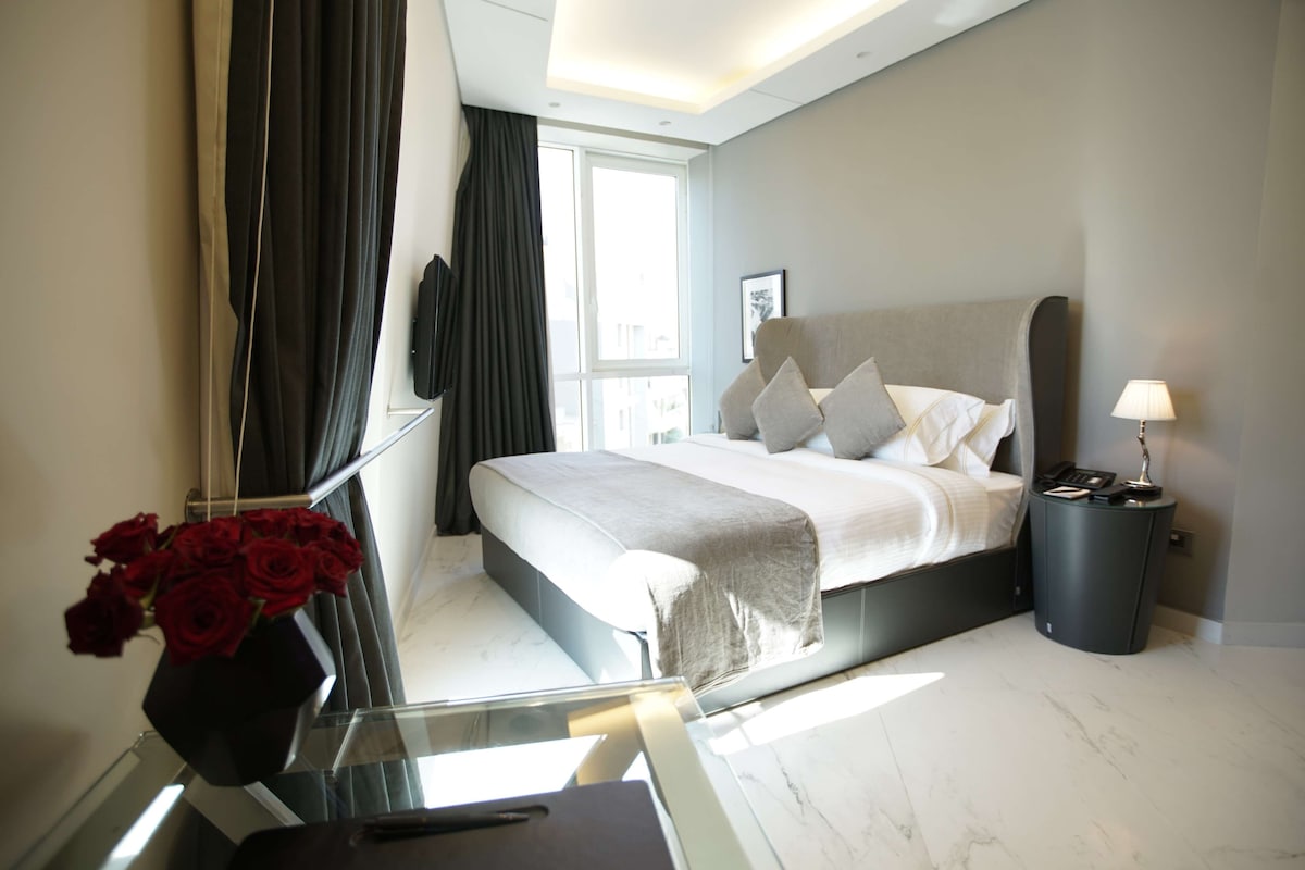 A modern hotel room with a large bed, a bedside table with a lamp, a mirror, and a vase with red roses.