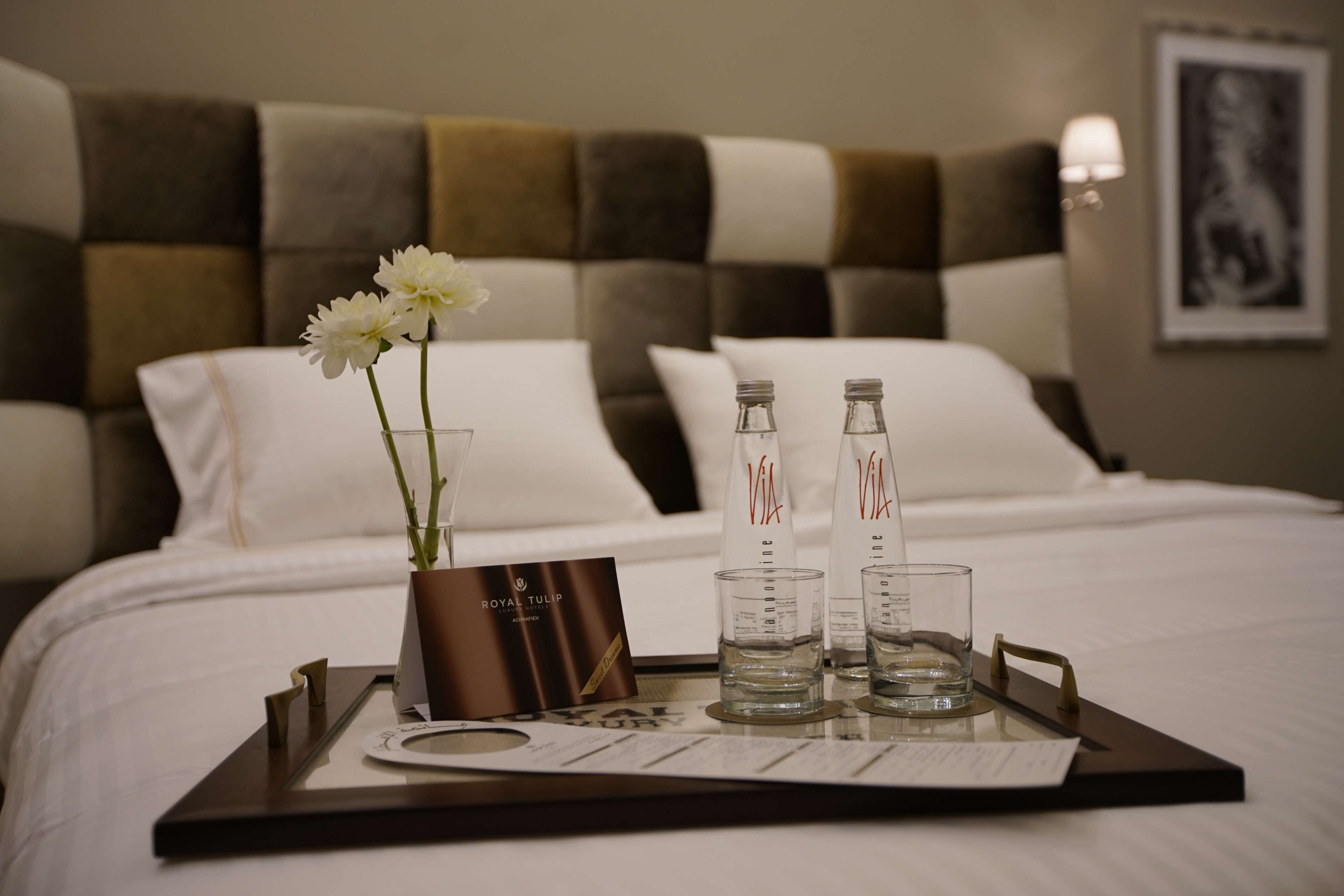 A neatly made bed with white linens, a tray with a vase of flowers, two bottles of water, and a card on a tray.