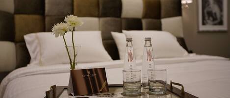 A neatly made bed with white linens, a tray with a vase of flowers, two bottles of water, and a card on a tray.