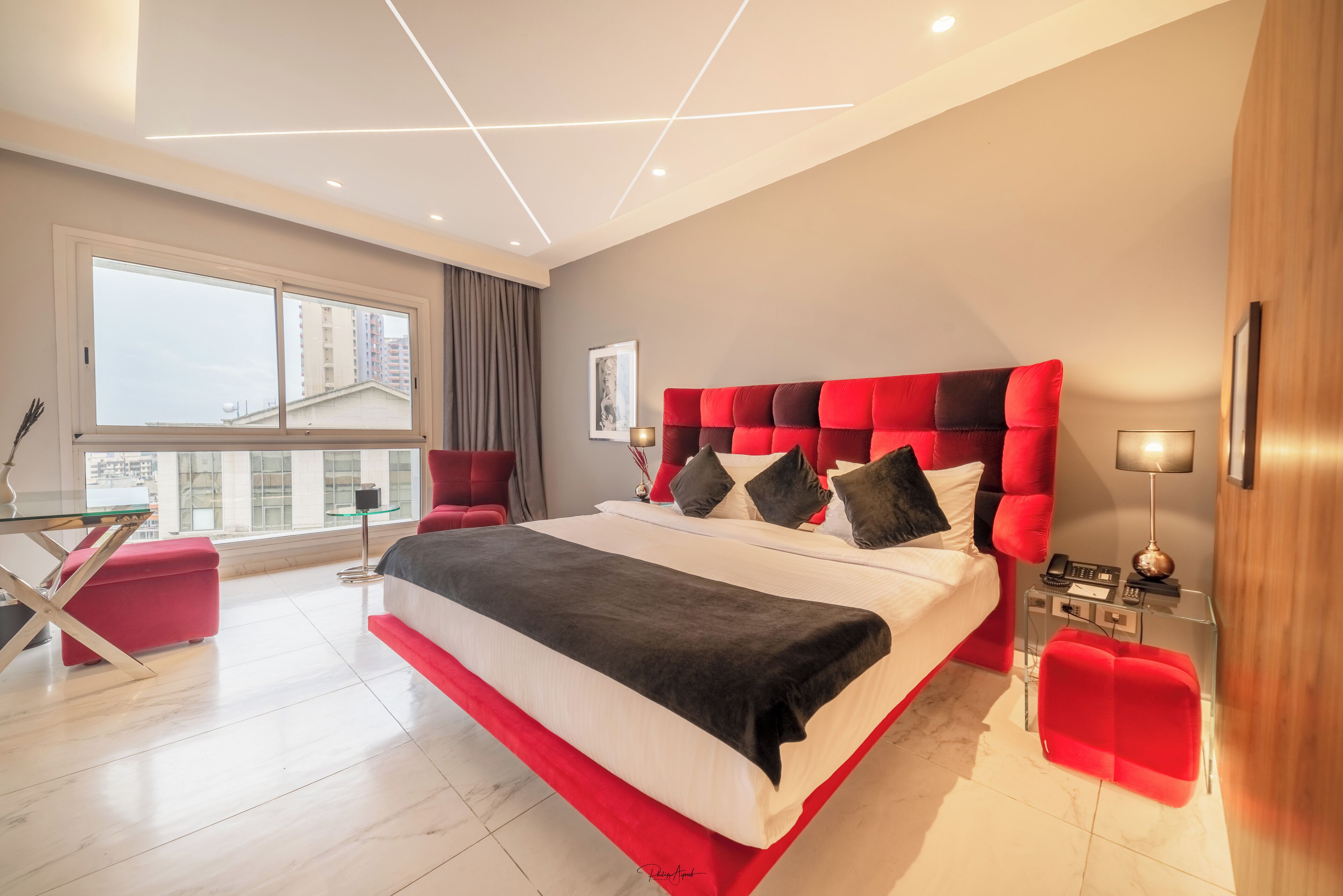 A modern bedroom with a large bed, a red upholstered headboard, a glass table, and a city view through the window.