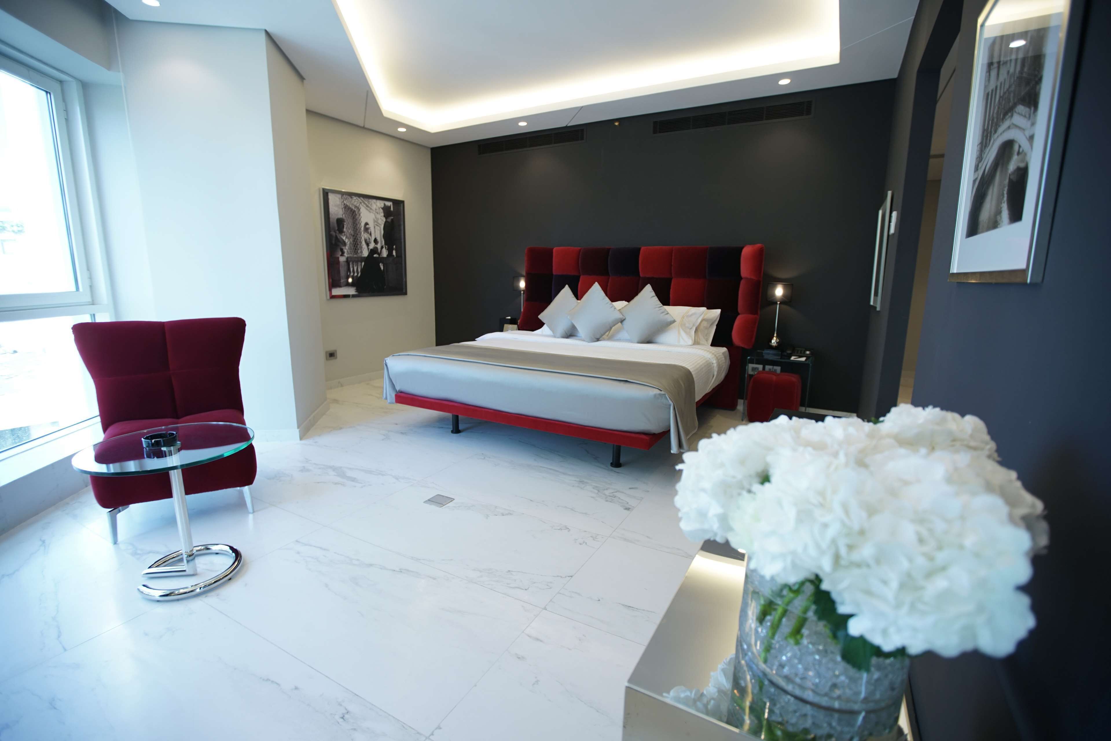 Une chambre à coucher moderne avec un lit, un fauteuil rouge, une table en verre et un vase de fleurs.