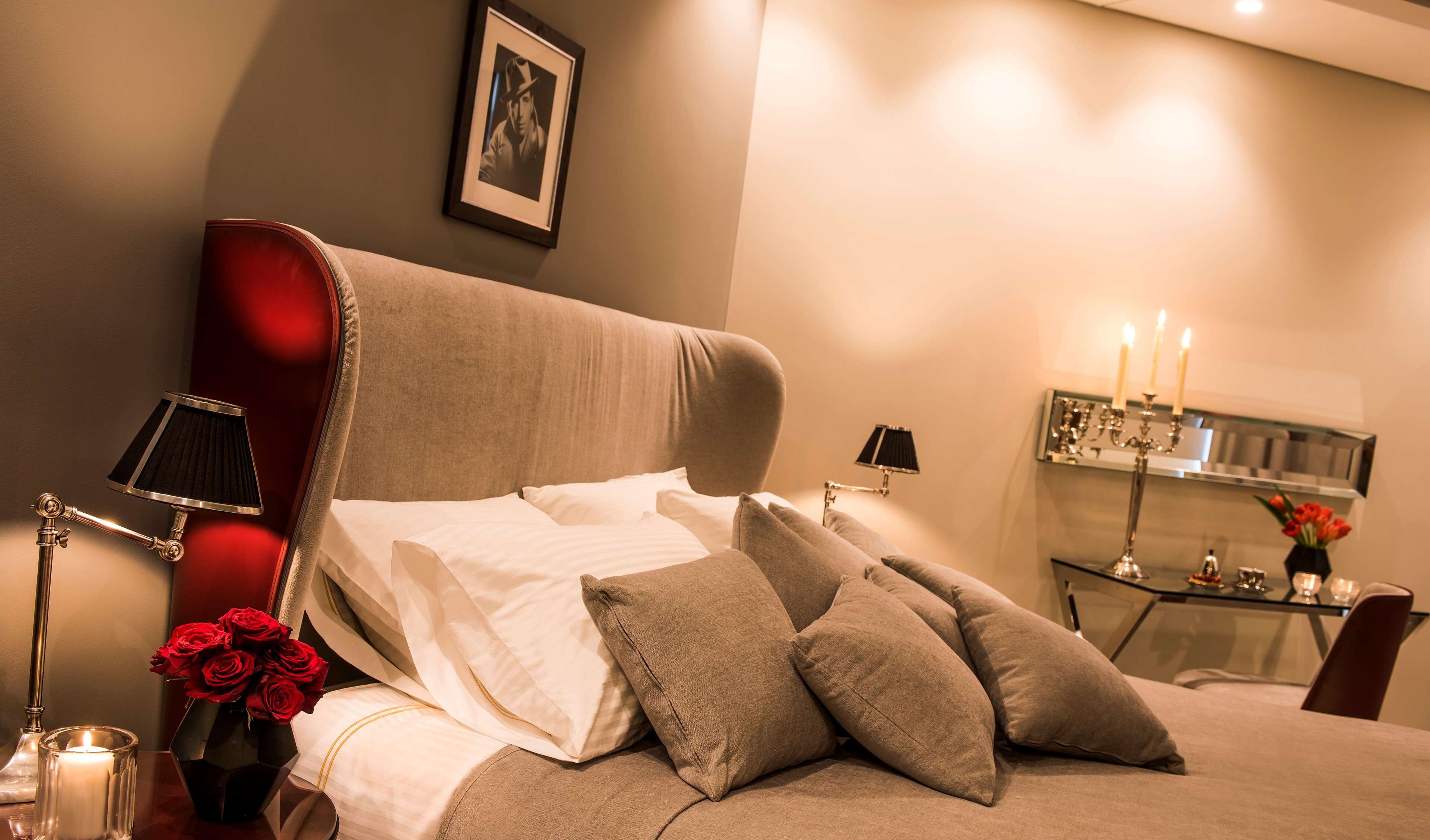 A neatly made bed with pillows, a bedside table with a lamp, a vase with red roses, and a framed picture on the wall.