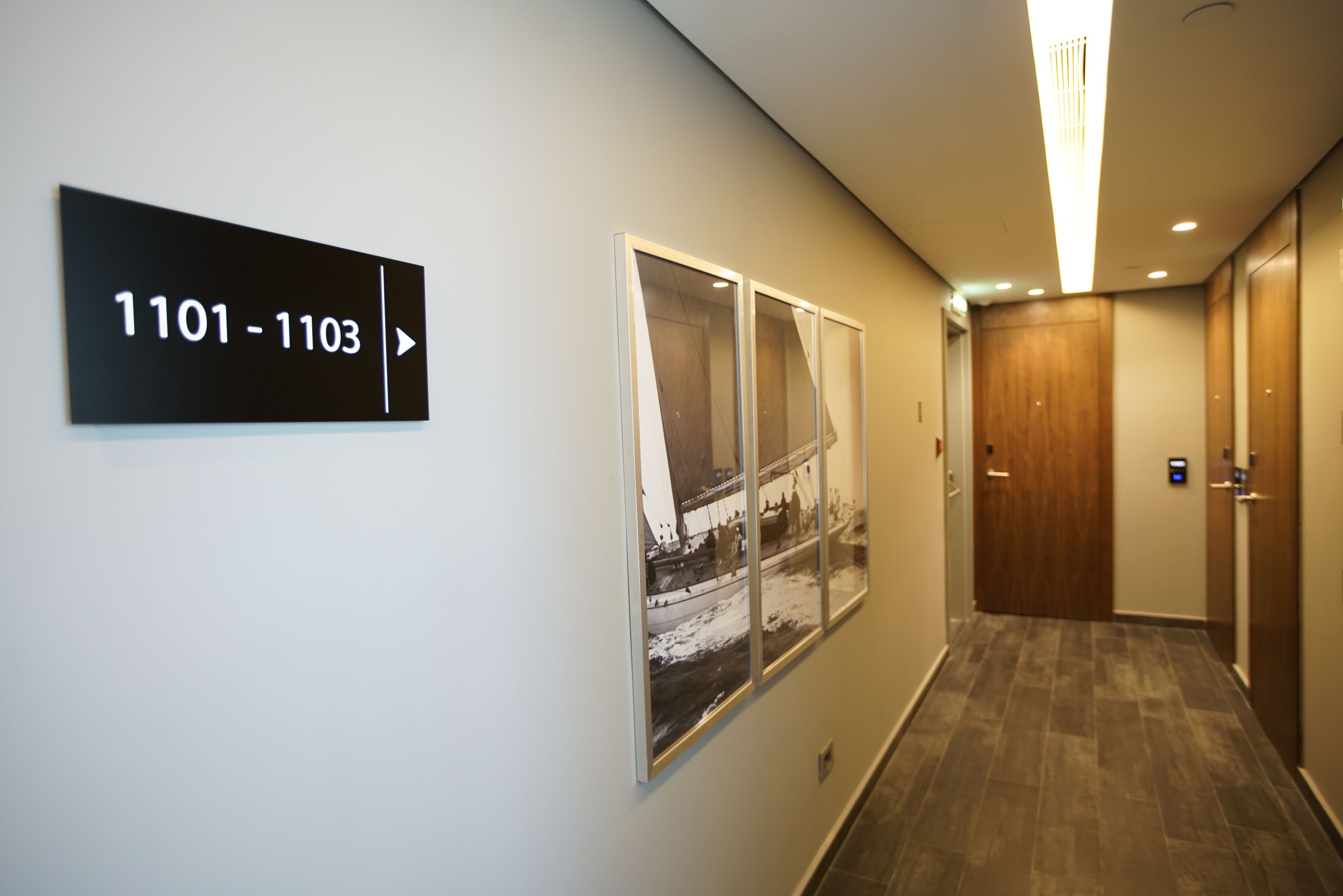 Un couloir avec un panneau indicateur pour les chambres 1101 à 1103, une grande photographie encadrée accrochée au mur et une porte en bois au bout du couloir.
