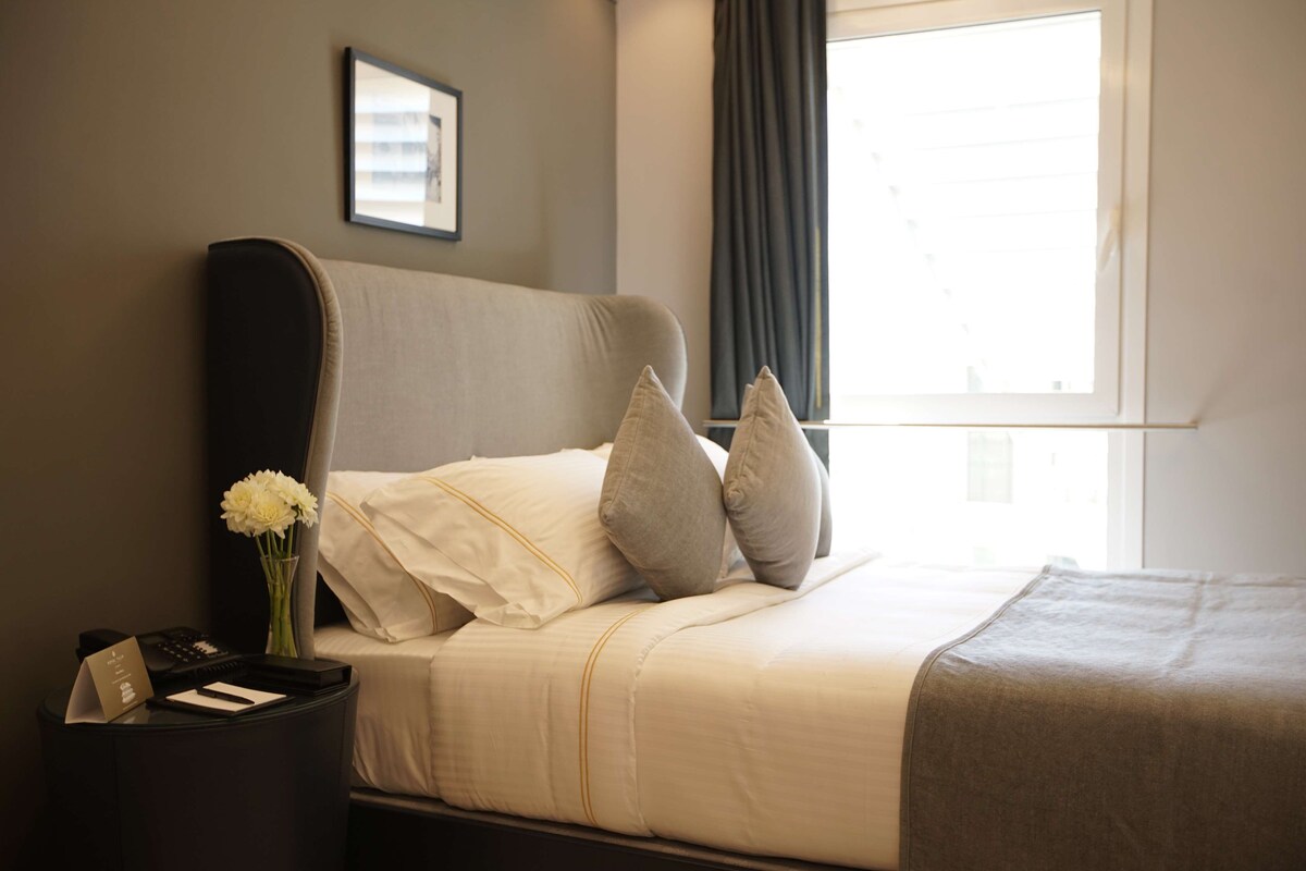 A neatly made bed with pillows, a bedside table with a vase of flowers, and a window with curtains.