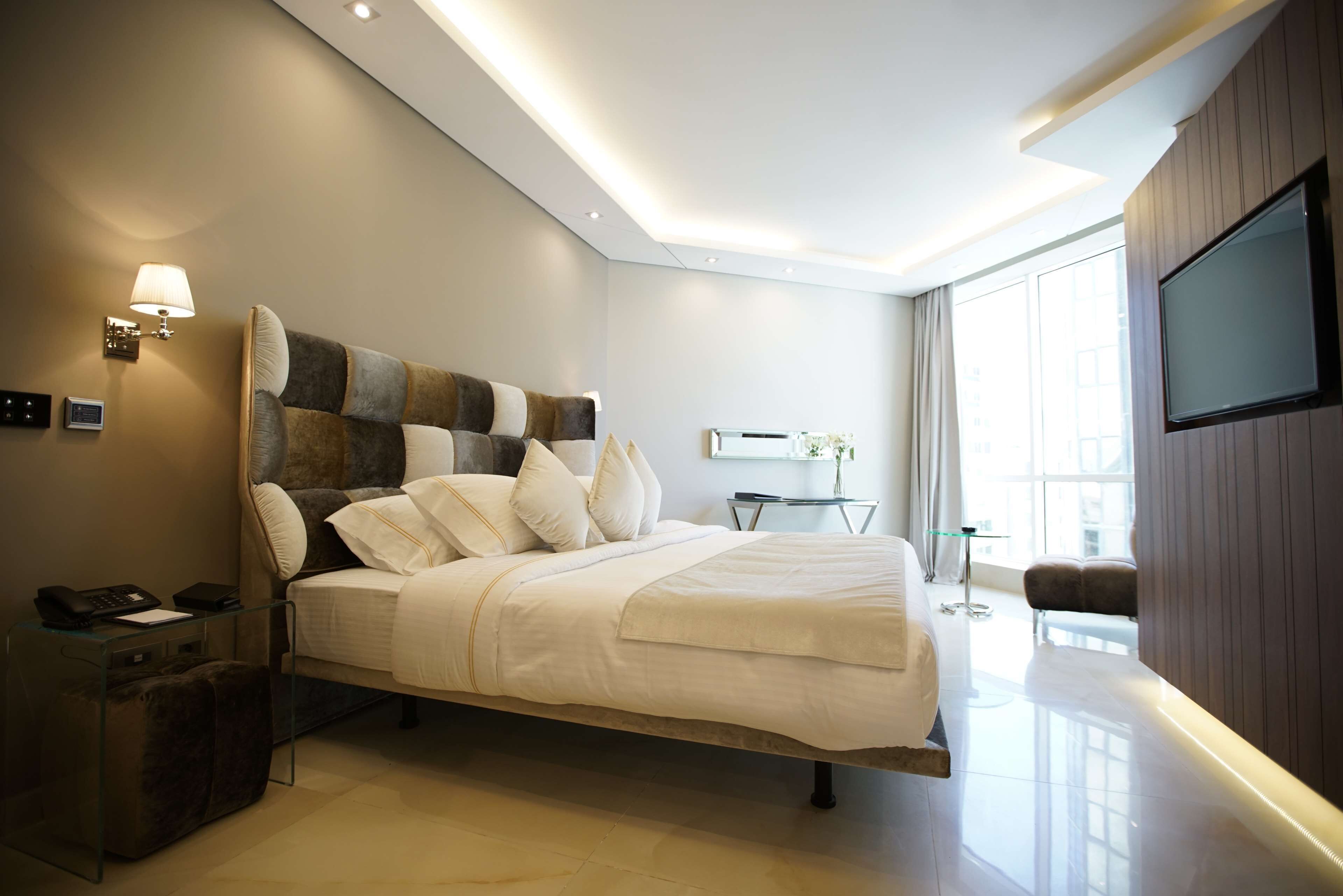 Un hotel moderno con una cama grande, un televisor de pantalla plana, una mesita de noche con lámpara y vistas a la ciudad a través de amplias ventanas.