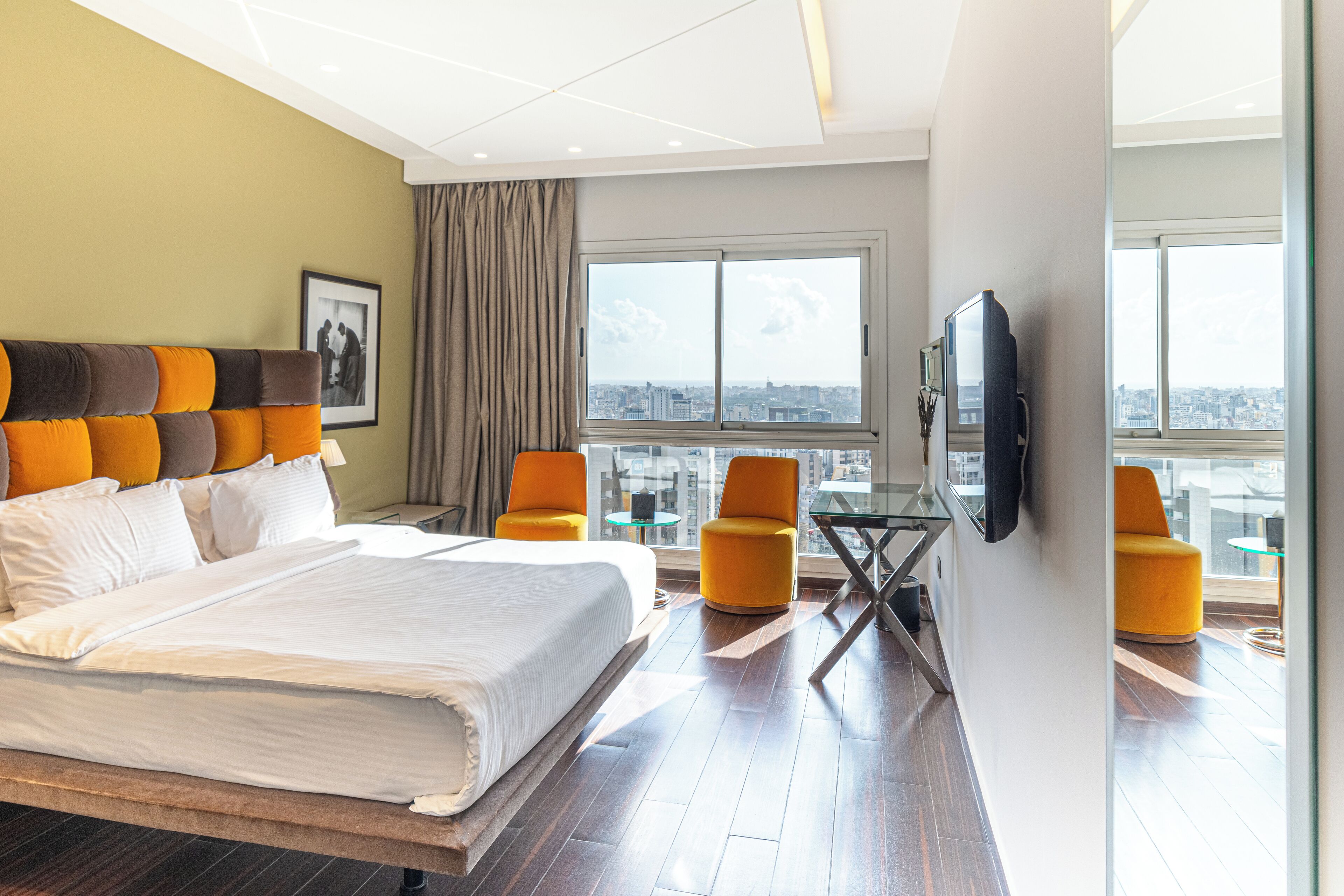 A hotel room with a large bed, two yellow armchairs, a glass table, a TV mounted on the wall, and a city view through large windows.
