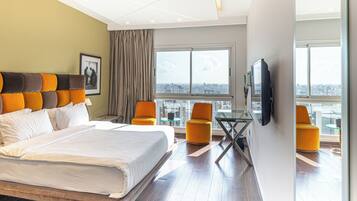 A hotel room with a large bed, two yellow armchairs, a glass table, a TV mounted on the wall, and a city view through large windows.