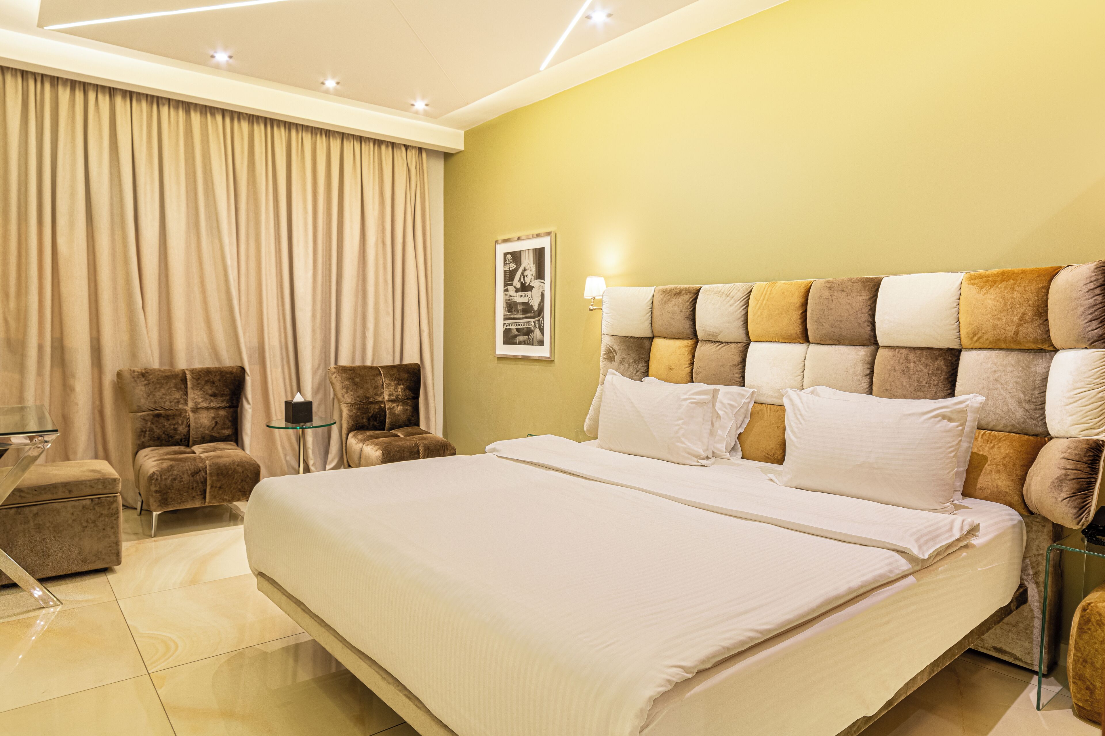 A modern hotel room with a large bed, a sitting area with a chair and a small table, and a large window with curtains.