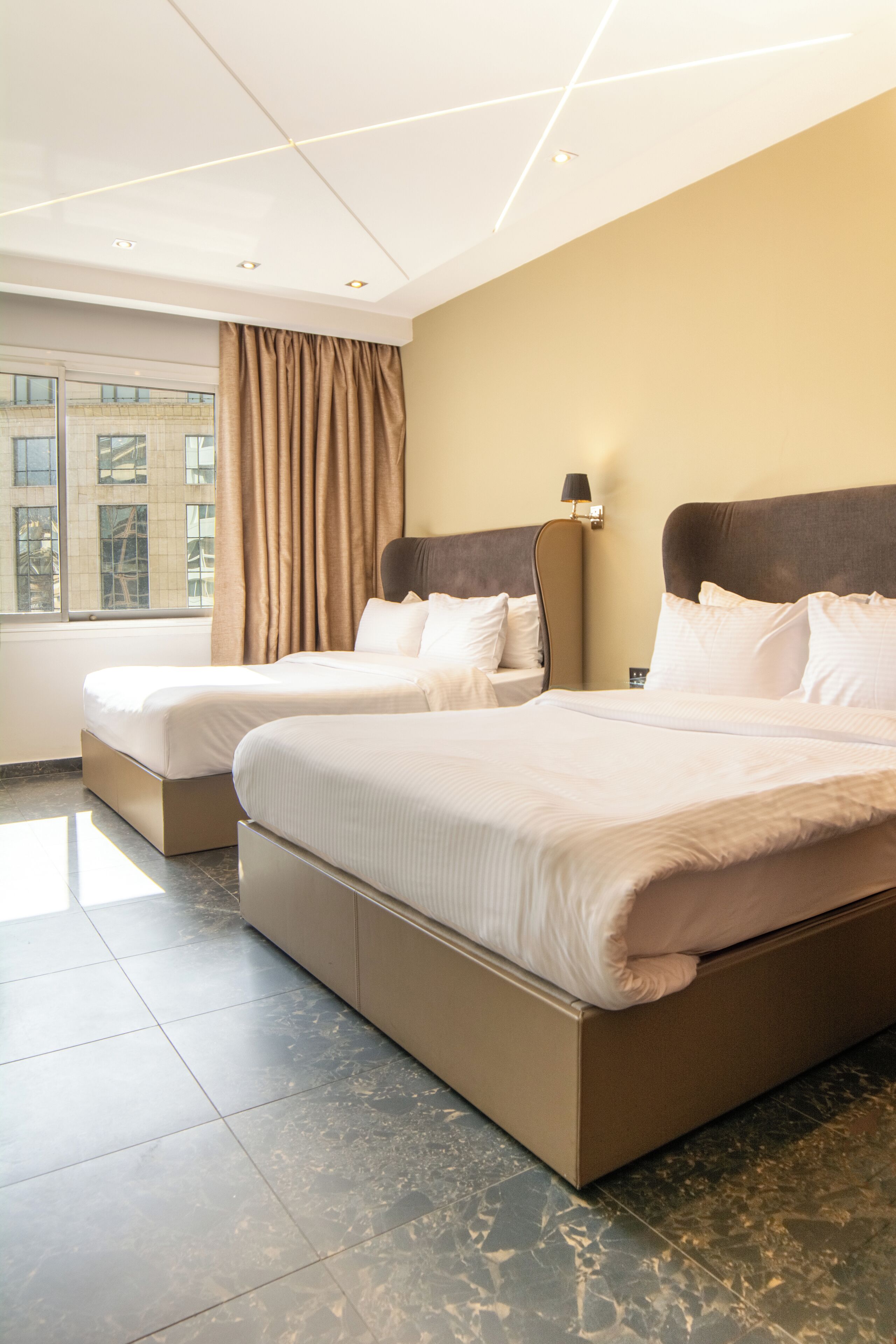 Habitación de hotel con dos camas, un ventanal con cortinas, una lámpara de pared y suelo de baldosas.