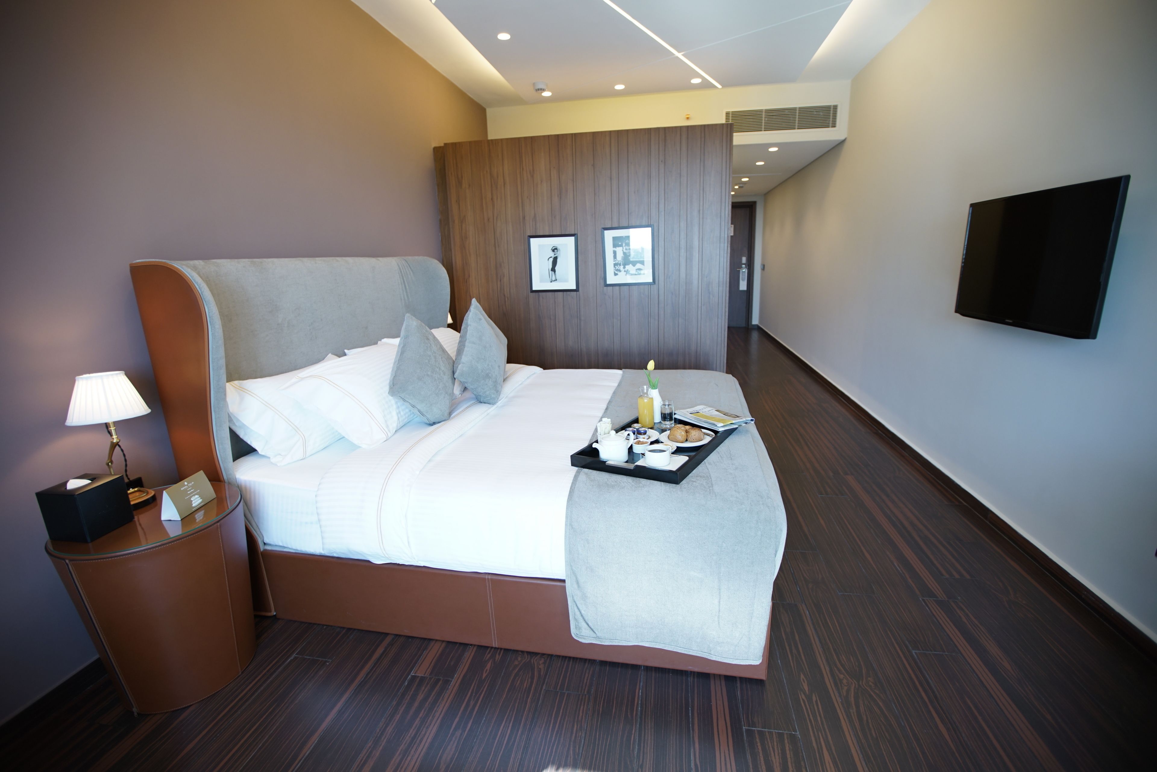 A modern hotel room with a large bed, a bedside table with a lamp, a tray with breakfast items, and a flat-screen TV on the wall.