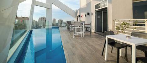 A rooftop pool area with a glass barrier, outdoor seating, and a city view.