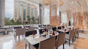 A modern dining area with tables set for service, large windows offering a view of a building, and decorative hanging lights. - Royal Tulip Achrafieh (Beirut)